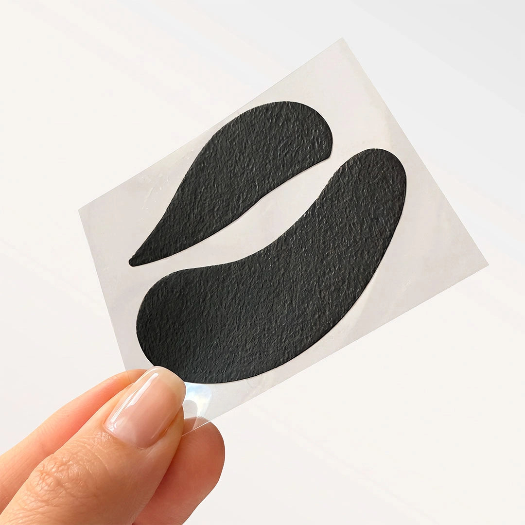 Une main tenant une feuille de support transparente avec une paire de patchs noirs bio-gel pour les paupières supérieures et le contour inférieur des yeux.